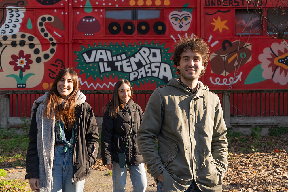 Tre giovani sorridenti in primo pianoe nello sfondo un muro con la scritta "Vai il tempo passa!