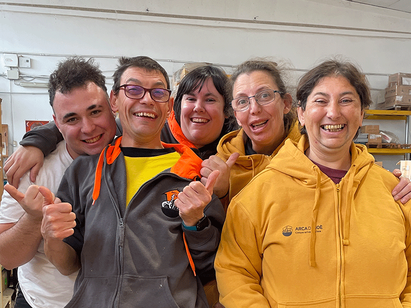 Un gruppo di cinque persone con disabilità sorridenti, abbracciate in un ambiente lavorativo, indossano abiti casual e felpe colorate. Sullo sfondo scaffali con materiali.