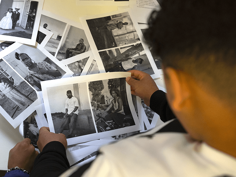 Una persona osserva diverse fotografie in bianco e nero sparse su un tavolo; le immagini ritraggono principalmente ritratti di persone in ambienti urbani.